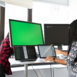 Two women working on laptops and monitors in a bright office setting, focused on technology and teamwork.